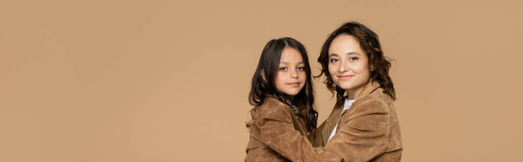 brunette woman and girl in trendy autumn jackets embracing while looking at camera isolated on beige, banner - Photo, Image