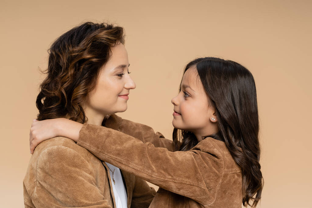 zijaanzicht van moeder en dochter in suède jassen glimlachend naar elkaar geïsoleerd op beige - Foto, afbeelding