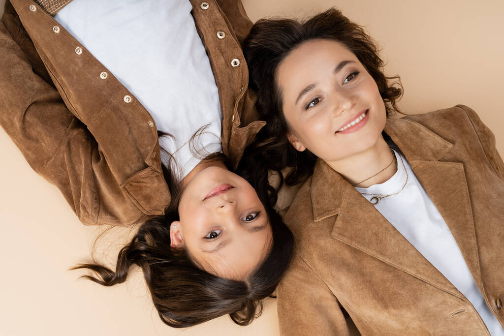 top view of brunette girl lying near mom in trendy autumn jacket and smiling at camera on beige background - Photo, Image