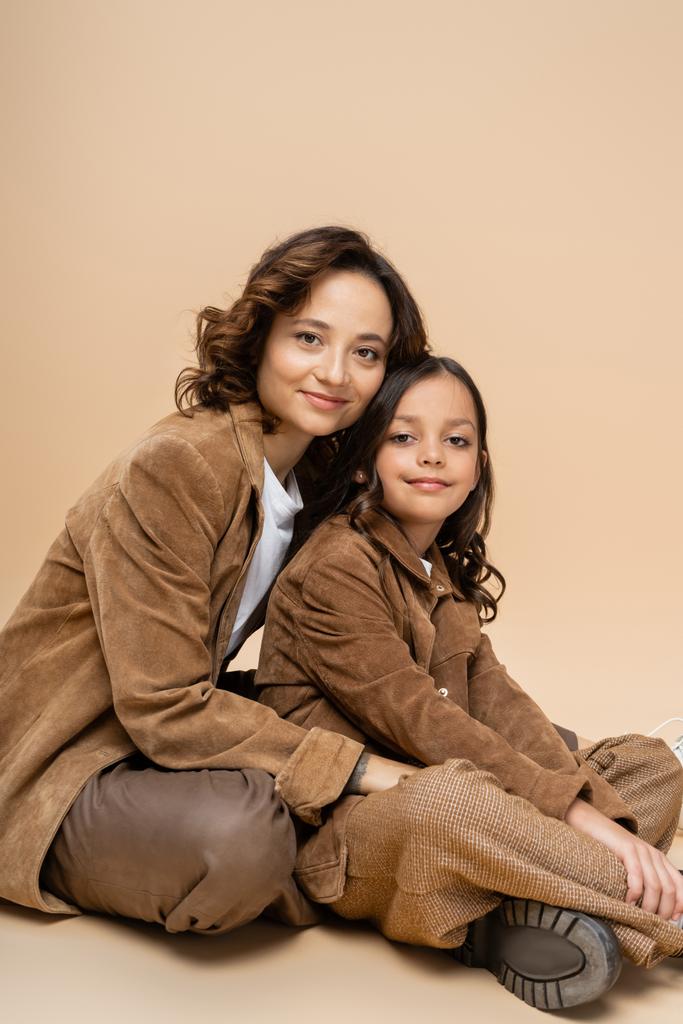 θετική μητέρα και παιδί σε suede μπουφάν και καφέ παντελόνι κάθεται και κοιτάζοντας κάμερα σε μπεζ φόντο - Φωτογραφία, εικόνα