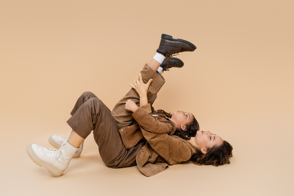 side view of girl with raised legs having fun and lying on mom in autumn outfit on beige background - Photo, Image