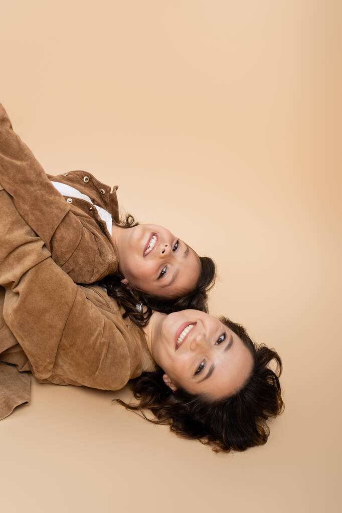 top view of cheerful woman and child in brown suede jackets lying and looking at camera on beige background - Photo, Image