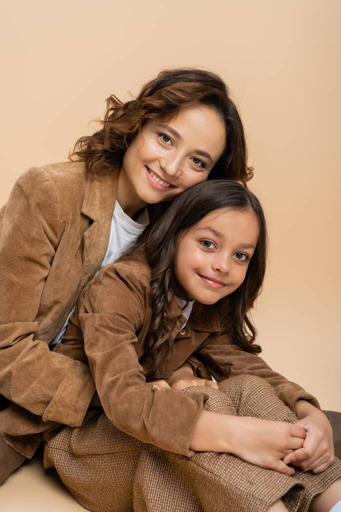 heureux mère et fille en daim marron vestes regardant caméra sur fond beige - Photo, image