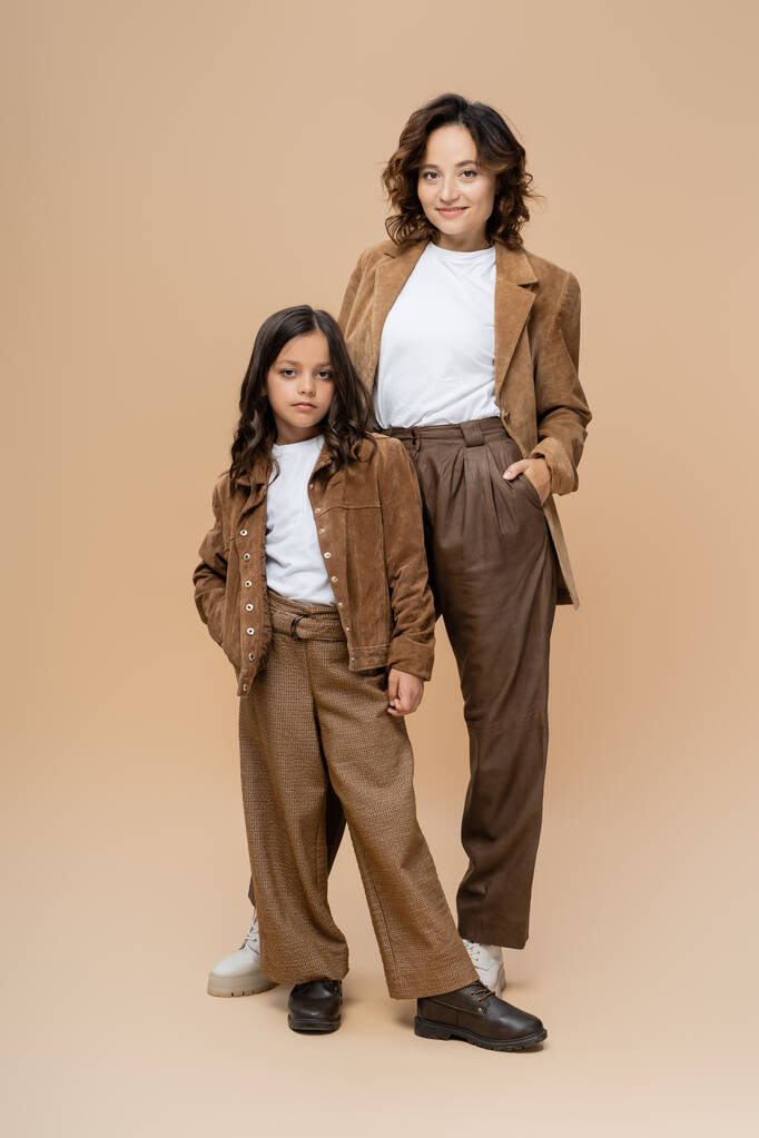 longitud completa de la mujer y la niña en ropa de otoño de moda posando con las manos en los bolsillos sobre fondo beige - Foto, imagen
