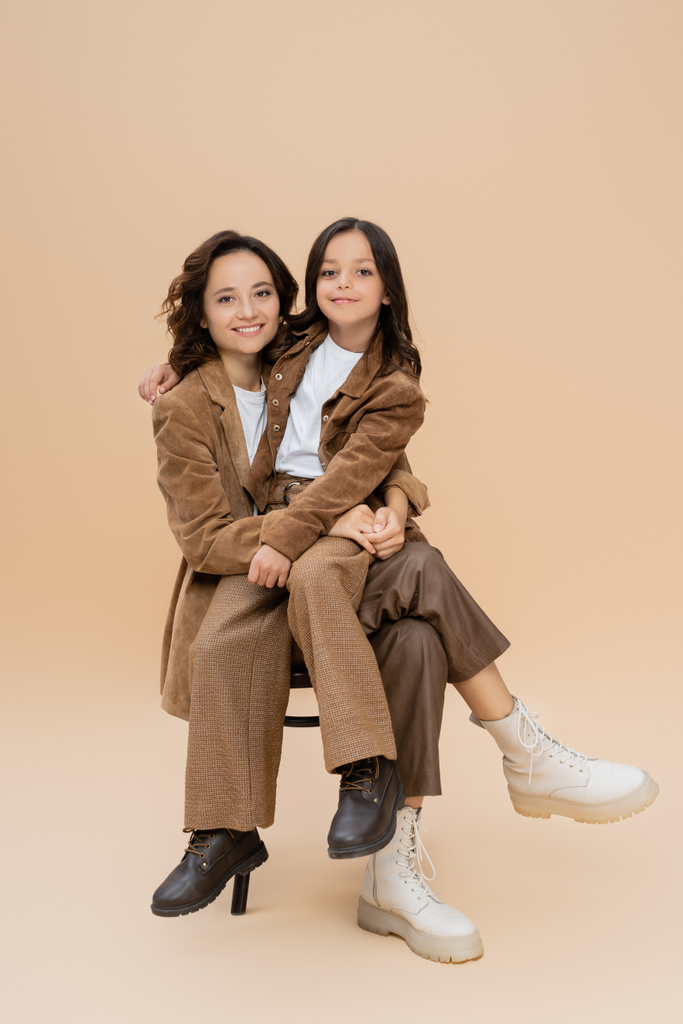 happy mother and daughter in trendy autumn outfit and boots sitting on chair on beige  - Photo, Image