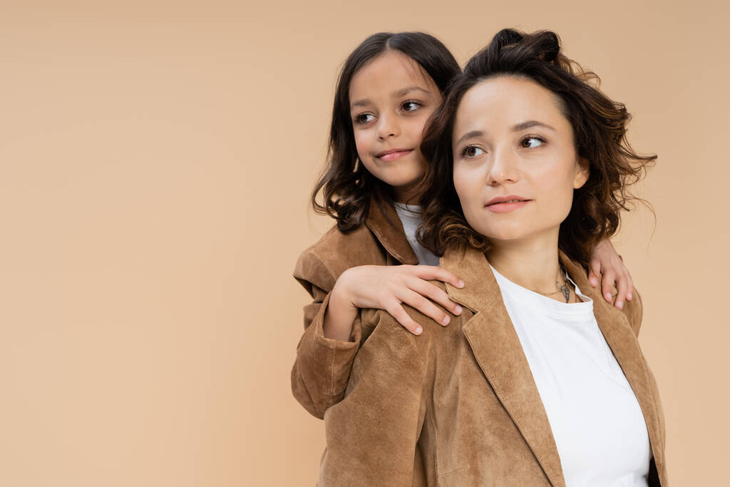 bruna ragazza abbracciare spalle di mamma in camoscio marrone giacca mentre guardando lontano isolato su beige - Foto, immagini