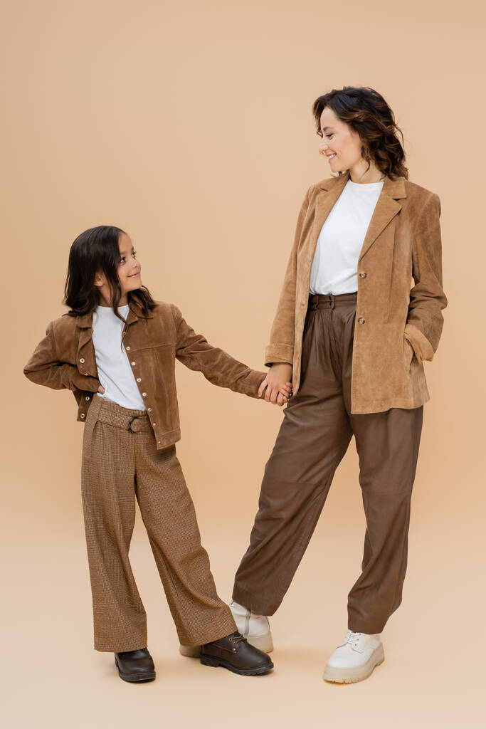 повна довжина жінки і дівчини в модному осінньому вбранні, тримаючись за руки і посміхаючись один одному на бежевому фоні
 - Фото, зображення