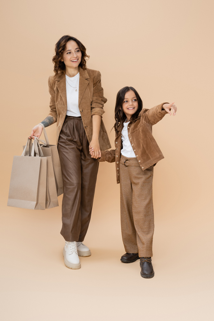 Lächelndes Mädchen hält Händchen mit trendiger Mutter mit Einkaufstüten und zeigt mit dem Finger auf beige  - Foto, Bild