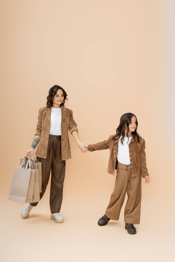 pleine longueur de fille en vêtements à la mode tenant la main avec maman heureuse avec des sacs à provisions sur fond beige - Photo, image