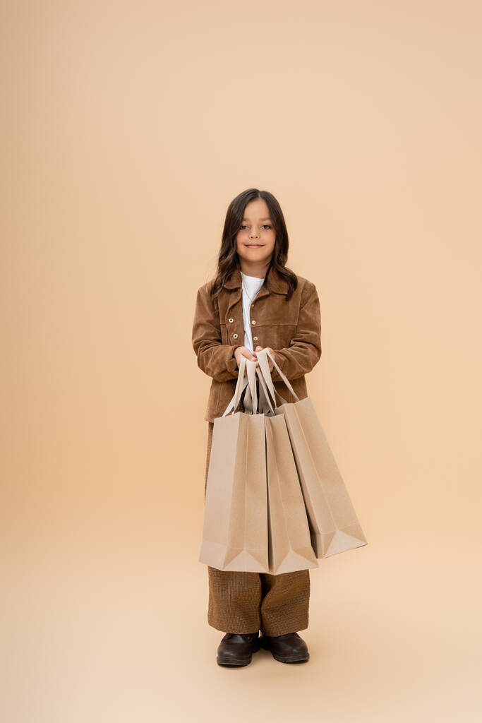 menina sorridente na moda roupas de outono segurando sacos de compras e olhando para a câmera no fundo bege - Foto, Imagem