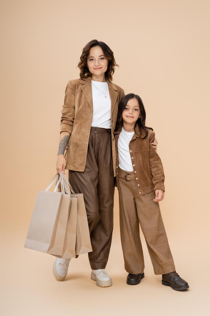 Frau in voller Länge mit Einkaufstaschen umarmt Tochter, während sie im trendigen Herbst-Outfit auf beige posiert  - Foto, Bild