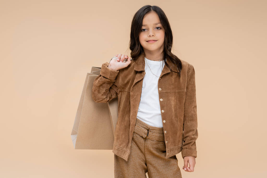 fille brune avec des sacs à provisions posant dans une veste en daim et regardant la caméra isolée sur beige - Photo, image