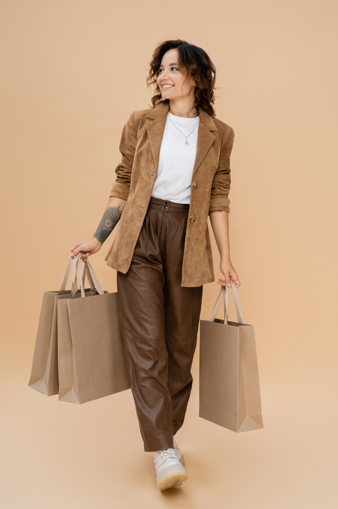 radosna kobieta w zamszowej kurtce spacerująca z torbami na zakupy i odwracająca wzrok na beżowym tle - Zdjęcie, obraz