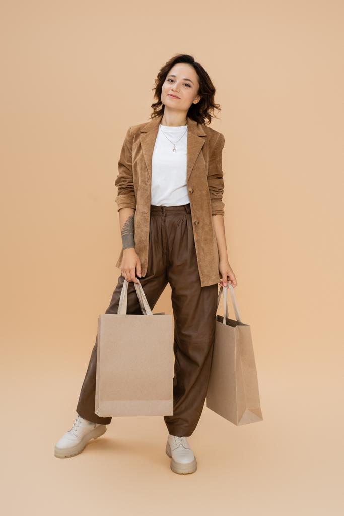 Güz kıyafetli, bej arka planda alışveriş torbalarıyla poz veren bir kadın. - Fotoğraf, Görsel