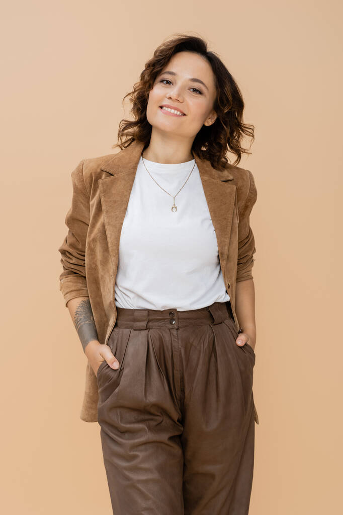 młoda kobieta w zamszowej kurtce stojąca z rękami w kieszeniach brązowych spodni odizolowanych na beżu - Zdjęcie, obraz