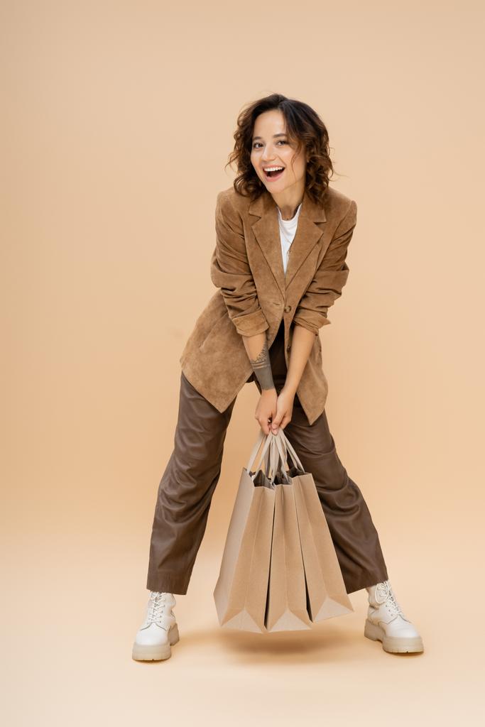 vzrušená žena s nákupními taškami při pohledu na kameru při pózování v módním podzimním oblečení na béžové  - Fotografie, Obrázek