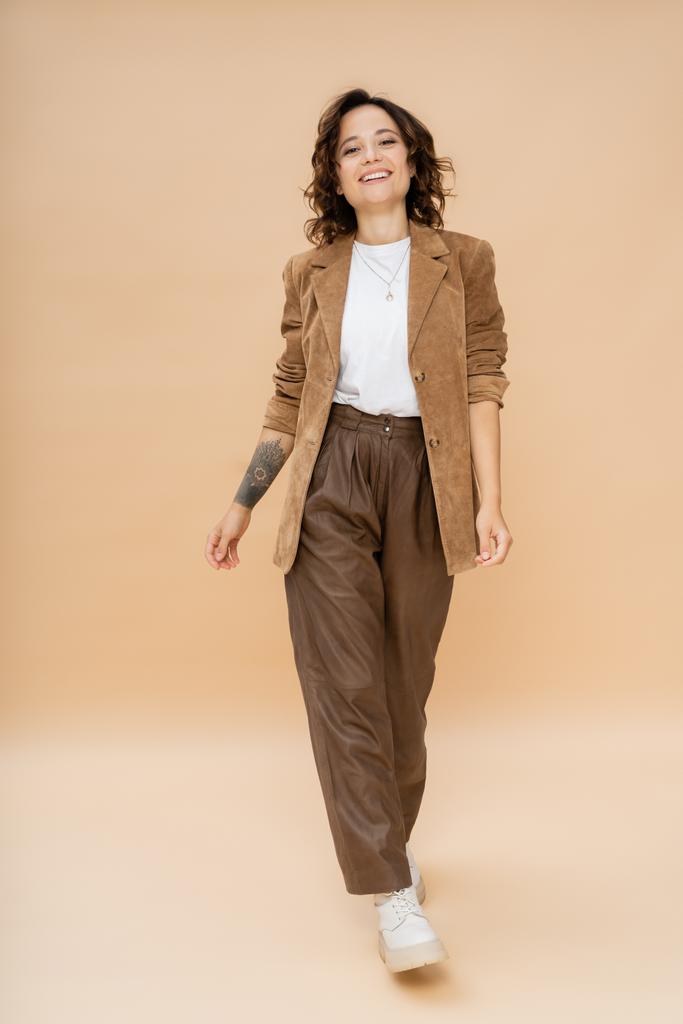 longitud completa de mujer tatuada en chaqueta de gamuza y pantalones marrones caminando sobre beige  - Foto, imagen