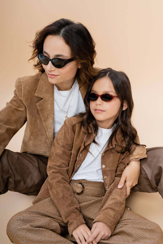 mère et fille à la mode dans des lunettes de soleil et vestes en daim marron assis sur fond beige - Photo, image
