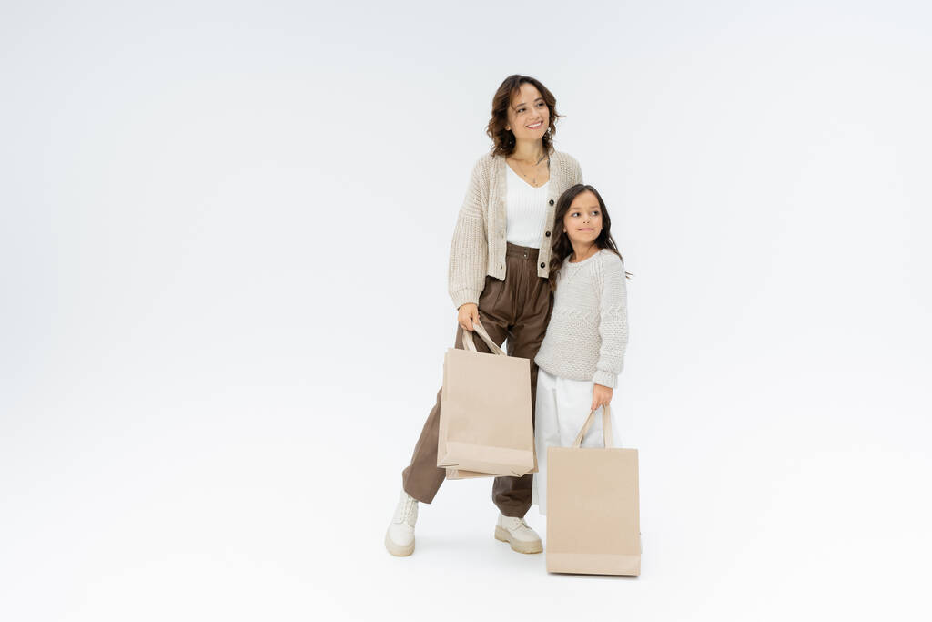 volledige lengte van trendy moeder en dochter met boodschappentassen wegkijkend op grijze achtergrond - Foto, afbeelding