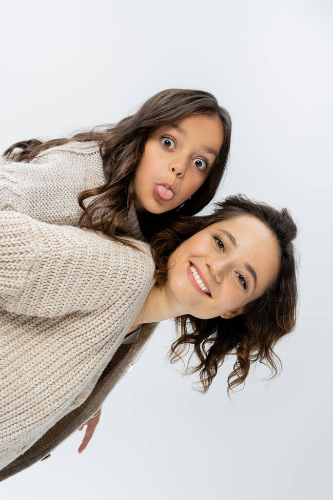 smiling woman in knitwear piggybacking daughter sticking out tongue isolated on grey - Photo, Image