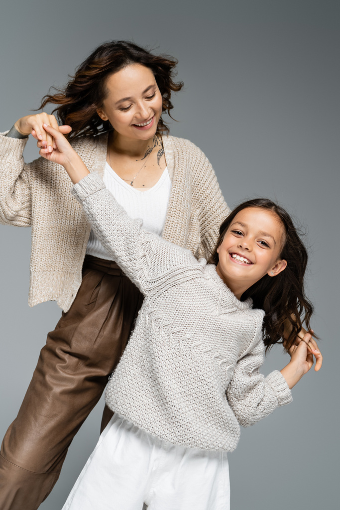 trendiges und fröhliches Mädchen blickt in die Kamera, während es mit glücklicher Mutter auf grau isoliert tanzt - Foto, Bild