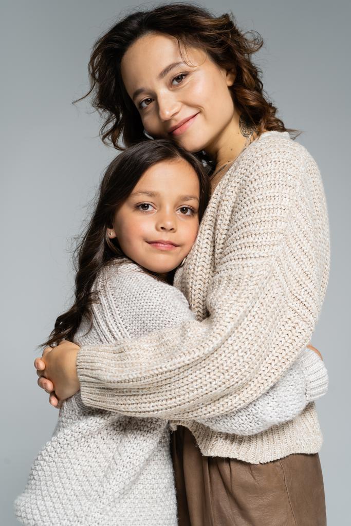 vrolijk mam en dochter in gezellig breigoed knuffelen en glimlachen op camera geïsoleerd op grijs - Foto, afbeelding