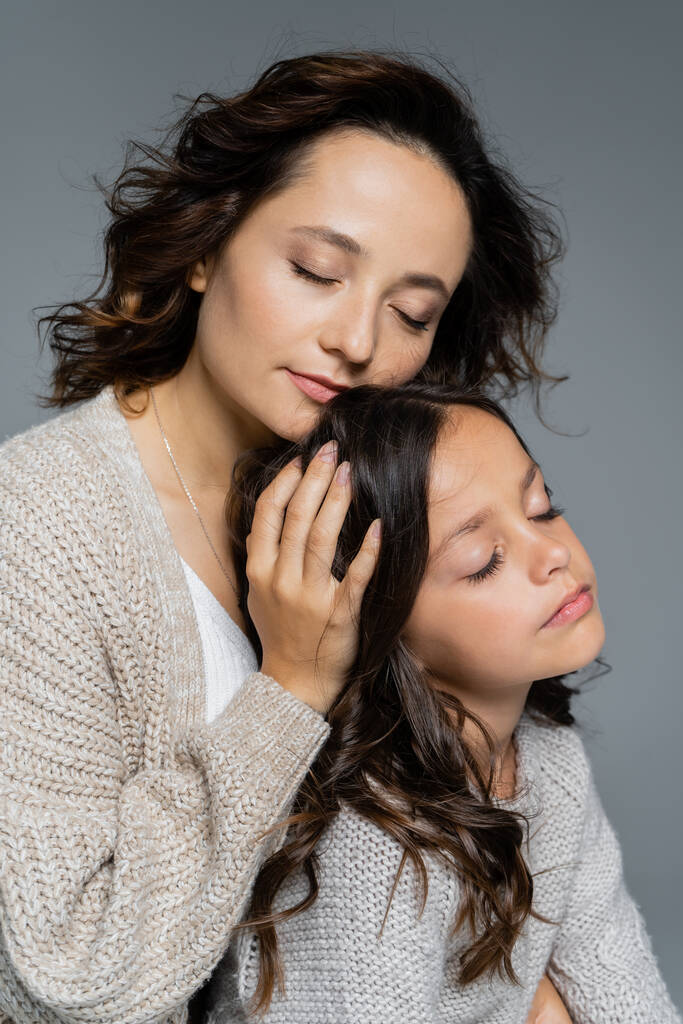 μελαχρινή γυναίκα με κλειστά μάτια αγκαλιάζει το κεφάλι της κόρης απομονώνονται σε γκρι - Φωτογραφία, εικόνα