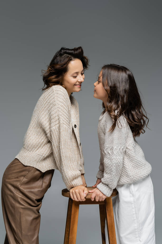 madre e hija de moda en prendas de punto cálidas mirándose cerca de taburetes de madera aislados en gris - Foto, imagen