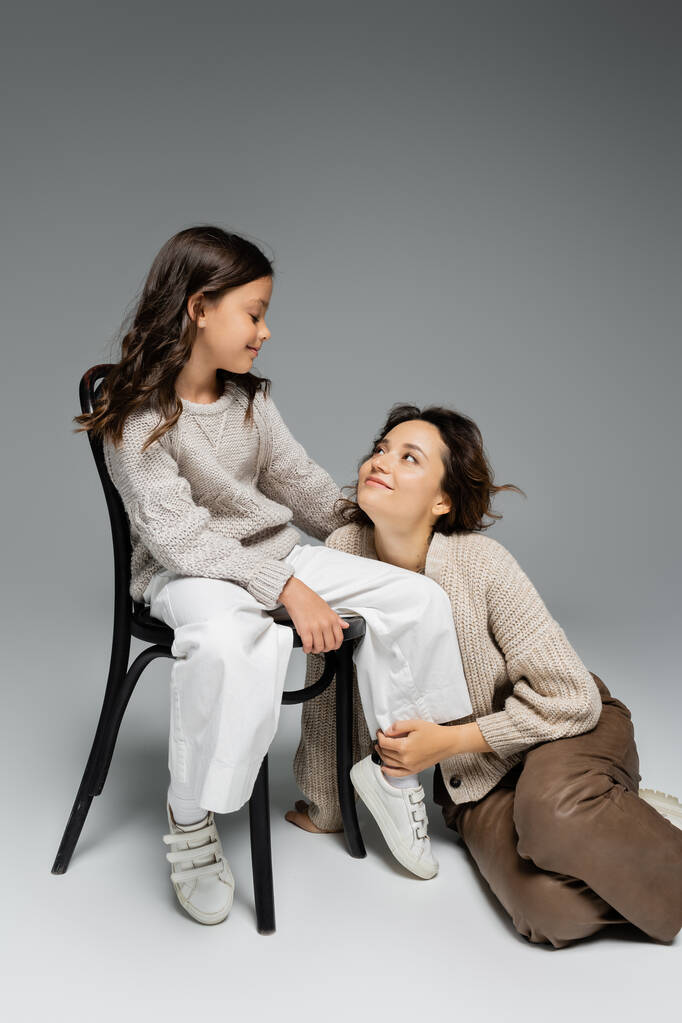 χαρούμενη γυναίκα κοιτάζοντας την κόρη κάθεται στην καρέκλα στο μοντέρνο φθινοπωρινό ντύσιμο σε γκρι φόντο - Φωτογραφία, εικόνα