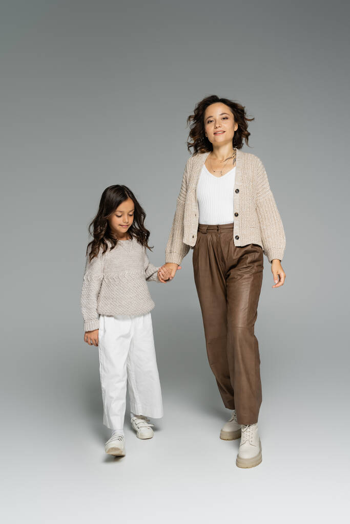 trendy woman smiling at camera while holding hands with daughter and walking on grey background - Photo, Image