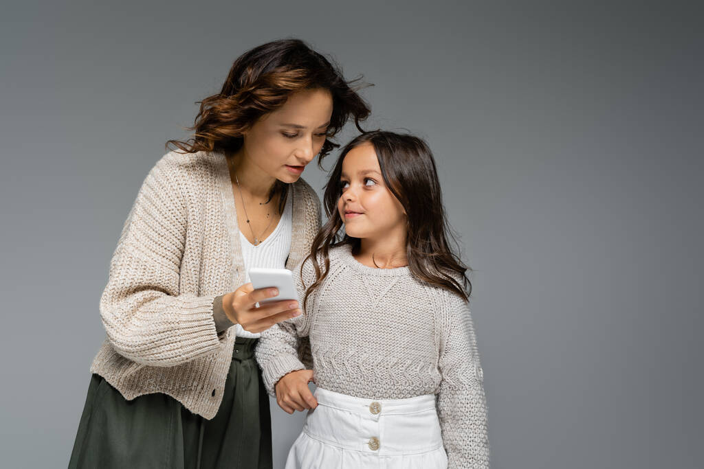 スタイリッシュな秋の服を着た笑顔の女の子は灰色で隔離されたスマートフォンでお母さんを見て - 写真・画像