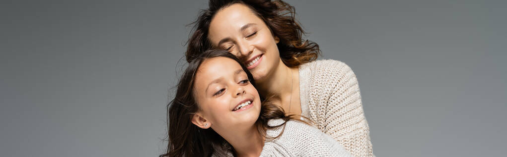 trendy vrouw glimlachen met gesloten ogen in de buurt gelukkig dochter geïsoleerd op grijs, banner - Foto, afbeelding