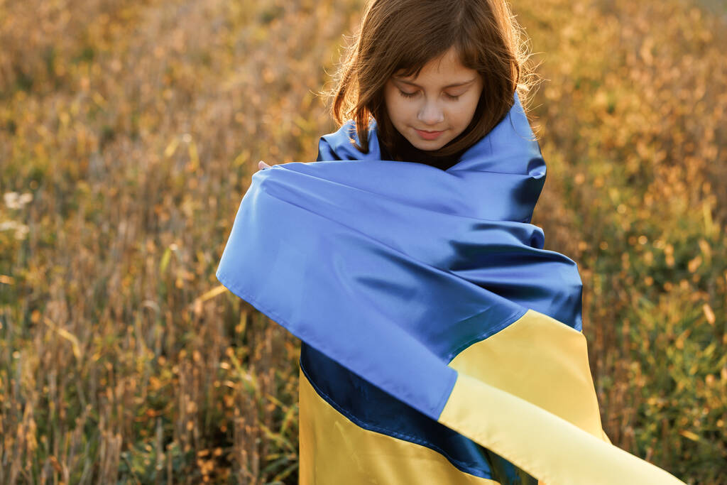 悲しいウクライナの少女の肖像画は、日没の空に対してフィールドの真ん中にウクライナのフラグで立っている。ウクライナの国旗を掲げる8歳の少女。ウクライナでの戦争。ウクライナへの添付 - 写真・画像