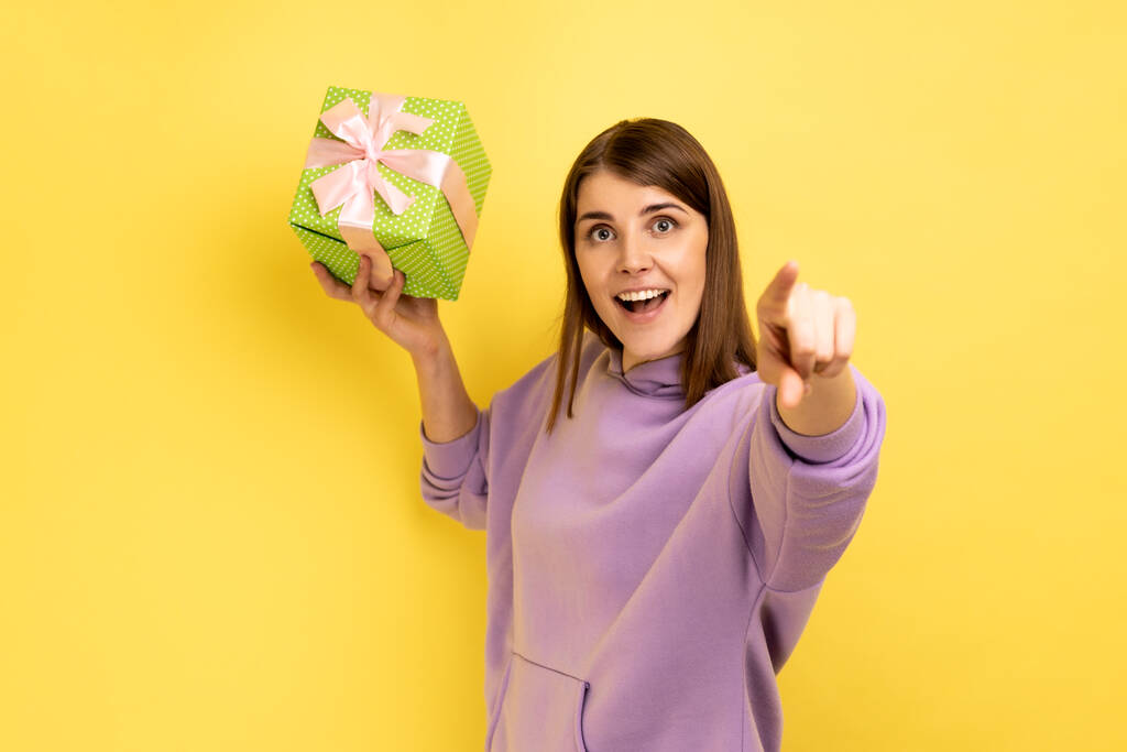 Szczęśliwa podekscytowana kobieta rzuca pudełko prezentów, mając szczęśliwy wygląd, świętując wakacje, gratulując i wskazując na aparat, ubrana w purpurowy kaptur. Kryty studio strzał izolowany na żółtym tle. - Zdjęcie, obraz