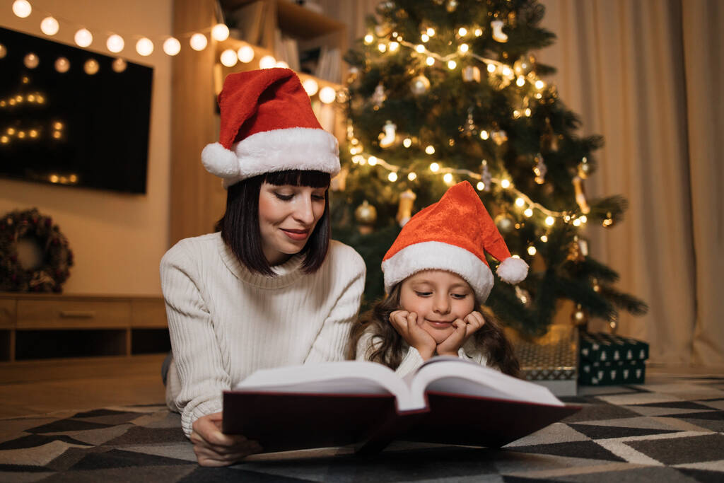 Bella giovane madre in cappello di Babbo Natale che legge libro di fiabe magiche a sua figlia sdraiata sul pavimento vicino all'albero di Natale, buon Natale. - Foto, immagini