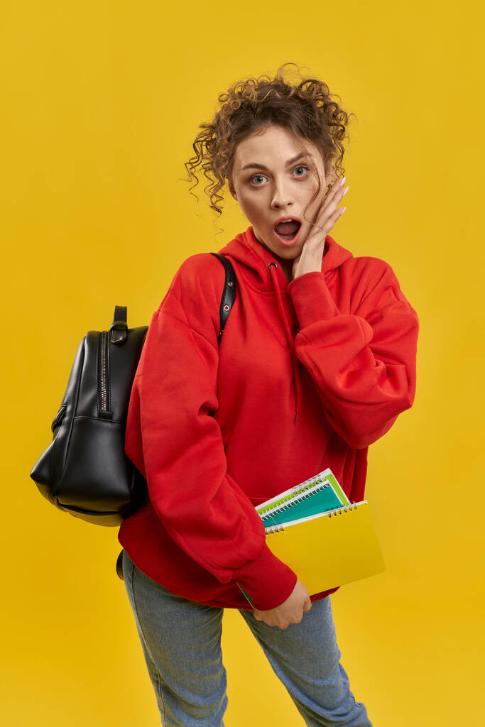 Front view of young student standing shocked, surprised with open mouth. Pretty woman with rucksack, holding books by hands, looking at camera. Isolated on yellow background. - Photo, Image