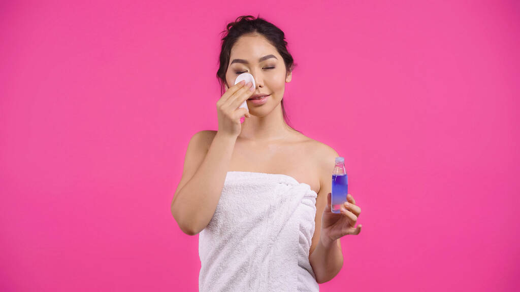 jeune femme asiatique en serviette tenant coton tampon avec de l'eau micellaire tout en nettoyant le visage isolé sur rose  - Photo, image