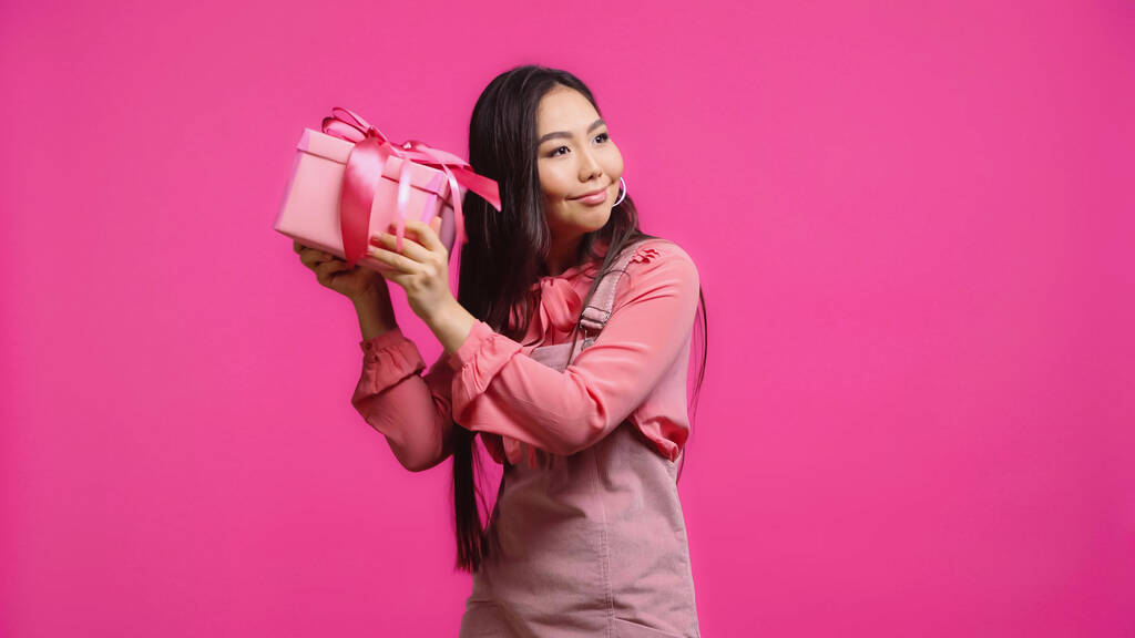 szczęśliwy młody Azji kobieta wstrząsające opakowanie prezent odizolowany na różowy  - Zdjęcie, obraz