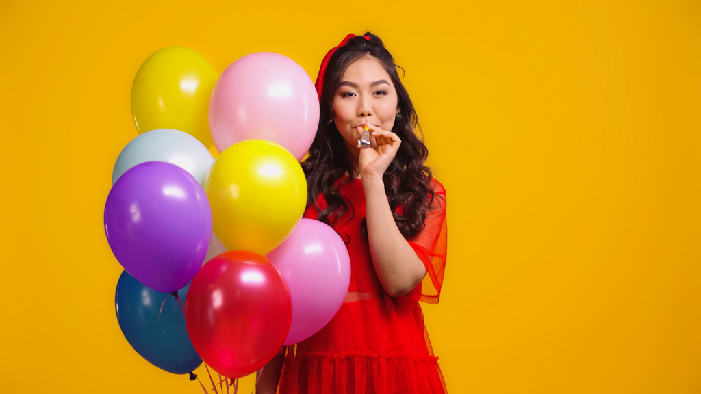 asiatische Frau in rotem Kleid mit bunten Luftballons und Blasen Party Horn isoliert auf gelb  - Foto, Bild