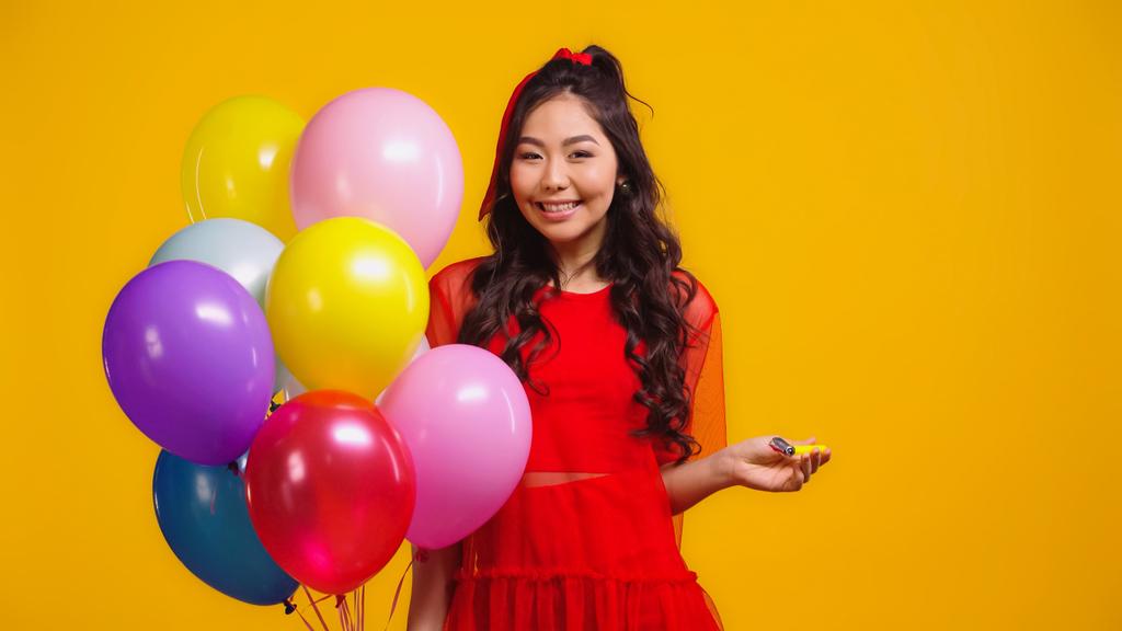 Kırmızı elbiseli neşeli Asyalı kadın elinde renkli balonlar ve sarıda izole edilmiş parti kornası tutuyor.  - Fotoğraf, Görsel
