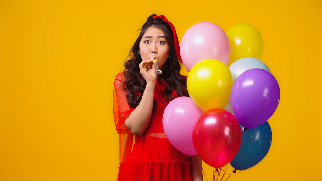 Asiatin im roten Kleid hält Luftballons in der Hand und bläst Partyhorn auf gelb  - Foto, Bild