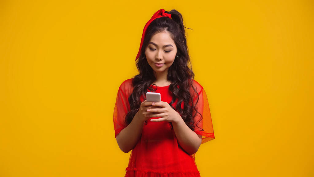 ευχαριστημένη Ασιάτισσα γυναίκα με κόκκινο φόρεμα γραπτών μηνυμάτων σε smartphone απομονωμένο σε κίτρινο  - Φωτογραφία, εικόνα