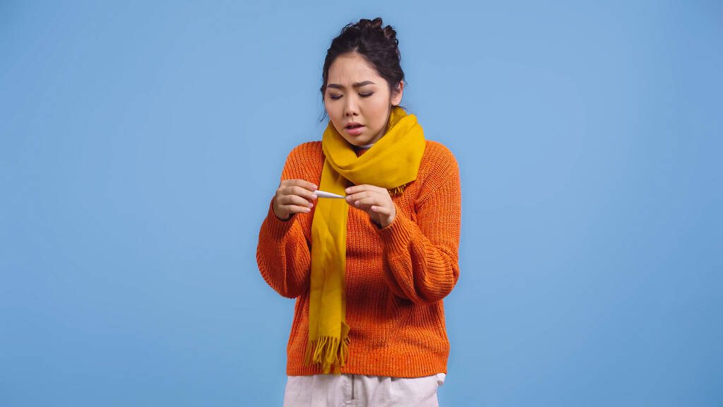 больная азиатка в свитере и шарфе с цифровым термометром, изолированным на голубом  - Фото, изображение