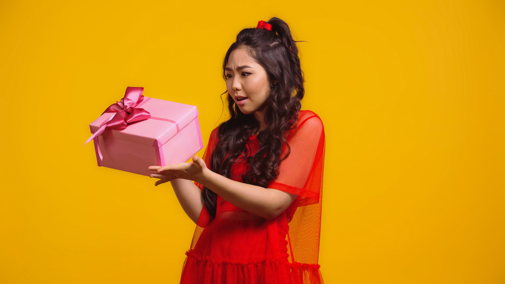 mujer asiática confundida sosteniendo regalo envuelto aislado en amarillo  - Foto, imagen
