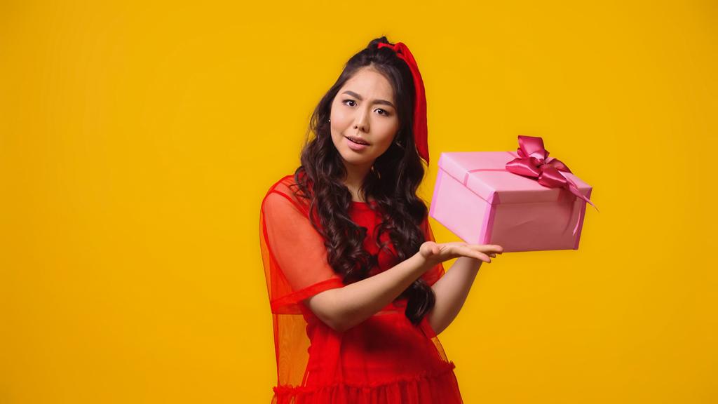 verwirrte junge asiatische Frau hält verpacktes Geschenk isoliert auf gelb  - Foto, Bild