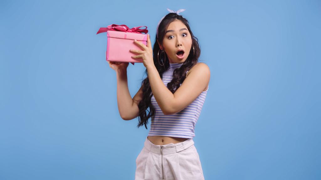 шокована молода азіатська жінка трясеться подарунок ізольовано на синьому
 - Фото, зображення
