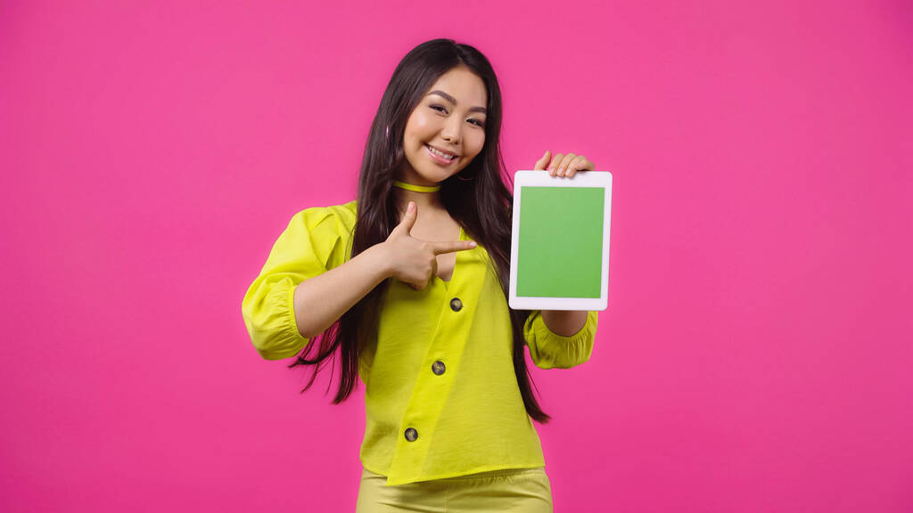 幸せなアジアの女性はピンクに隔離された緑の画面でデジタルタブレットを指しています  - 写真・画像