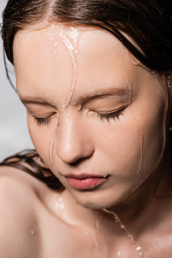 Zblízka pohled na vodu kapající na tvář mladé ženy izolované na šedé  - Fotografie, Obrázek