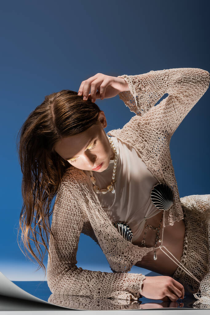Δίκαιη μαλλιά μοντέλο σε πλεκτή ζακέτα και μαργαριτάρι κολιέ κοιτάζοντας ανακλαστική επιφάνεια σε μπλε φόντο  - Φωτογραφία, εικόνα
