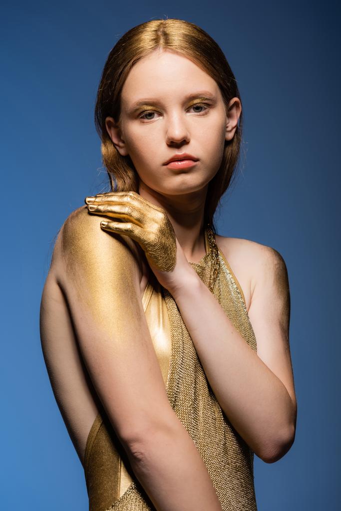 Retrato de modelo joven en vestido dorado y pintura mirando a la cámara aislada en azul  - Foto, Imagen